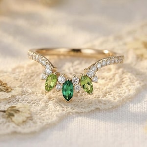 Puede incluir: Un anillo dorado con un diseño curvo, con una piedra preciosa verde esmeralda central y dos piedras preciosas verde claro a cada lado. El anillo está adornado con pequeñas piedras preciosas transparentes a lo largo de la banda. El anillo está sobre una superficie de encaje blanco.