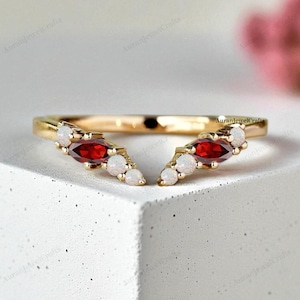 May include: A gold ring with a curved design. The ring features two marquise-cut red gemstones and several small, round, white gemstones. The ring is set on a white surface, with a blurred pink flower in the background.