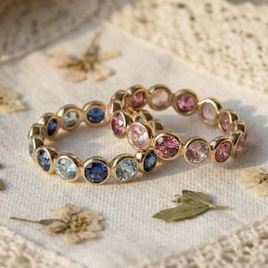 Peut inclure: Deux bagues en or serties de pierres précieuses colorées. L'une présente des pierres bleues et bleu clair, l'autre des pierres roses. Les bagues sont posées sur un tissu blanc avec des fleurs séchées et une bordure en dentelle.