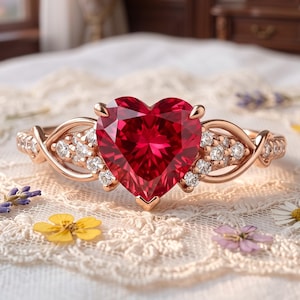 May include: A rose gold ring featuring a large, heart-shaped red gemstone at the center. The band is crafted with intricate, swirling metalwork and is accented with small, clear, round-cut stones along the sides, creating a detailed, elegant appearance.