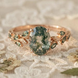 May include: Rose gold ring featuring an oval-cut moss agate gemstone. The ring has a leaf design with small green gemstones and diamond accents. The ring is set on a white lace surface.