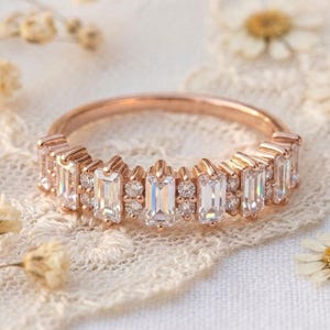 Op de afbeelding: Ros&eacute;gouden ring met een band van rechthoekige en rond geslepen heldere edelstenen. De ring ligt op een wit kanten oppervlak met kleine gedroogde bloemen.