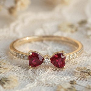 May include: A gold ring featuring a bow design with two heart-shaped, deep red gemstones and a small diamond in the center. The band is adorned with small diamonds. The ring is set against a soft, textured background.