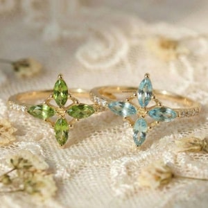 May include: Two gold rings, each with a gemstone flower design. One ring has green gemstones, the other blue. Both have delicate bands with small, clear stones. The rings are set against a soft, textured background.