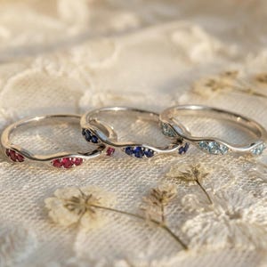 Może przedstawiać: Trzy srebrne pierścionki o falistym wzorze, każdy z rzędem małych kamieni szlachetnych. Jeden pierścionek ma czerwone kamienie, drugi niebieskie, a trzeci jasnoniebieskie. Pierścionki są prezentowane na białej koronkowej powierzchni.