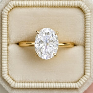 May include: A gold engagement ring featuring an oval-cut diamond. The ring is set with four prongs and presented in a cream-coloured ring box. The diamond appears to be approximately 2.5 cm in length.
