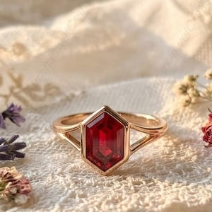 Puede incluir: Un anillo dorado con una gema hexagonal de color rojo intenso. El anillo está sobre una superficie de encaje blanco, con flores secas y ramitas de lavanda. El diseño del anillo presenta una caña dividida.