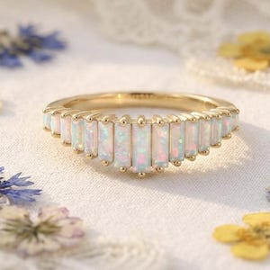 May include: A gold ring with a row of rectangular, iridescent opal stones. The ring is set against a white background with dried flowers. The opal stones have a range of colors, including white, pink, and blue.
