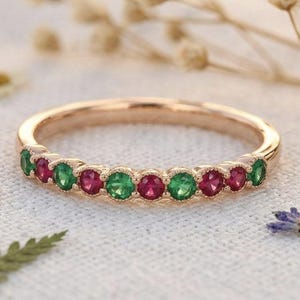 May include: Rose gold ring with alternating emerald and ruby gemstones. The ring features a delicate band and a row of vibrant green and red stones, creating a colorful and elegant design. The ring is displayed on a white surface with floral accents.
