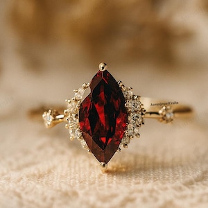 May include: A marquise-cut, deep red gemstone ring with a gold band. The gemstone is surrounded by smaller, clear stones. The ring is set on a cream-colored, textured surface. The ring is a piece of jewelry.