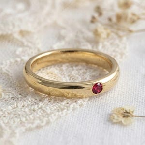 May include: A gold ring with a small, round, red gemstone set into the band. The ring is on a white lace surface, with dried flowers scattered around it. The ring is a simple band design.