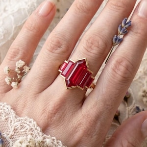 May include: A gold ring with a large, faceted, red gemstone. The ring has a geometric design with a central rectangular stone and two smaller stones on either side. The ring is displayed on a hand, with dried flowers and lace in the background.