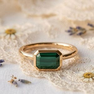 Może przedstawiać: Złoty pierścionek z prostokątnym, szmaragdowo-zielonym kamieniem szlachetnym. Pierścionek leży na delikatnej, kremowej koronkowej powierzchni, z małymi kwiatami i gałązkami lawendy. Obrączka pierścionka jest z prostego, polerowanego złota.