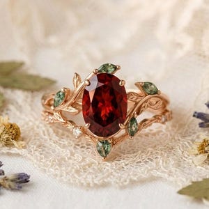 May include: A rose gold ring with a large oval-cut, deep red gemstone. The ring is decorated with delicate leaf and vine details, and smaller green gemstones. A smaller matching ring sits behind it.