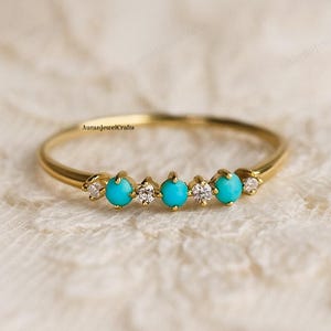 Op de afbeelding: Een delicate gouden ring met vijf turkooizen stenen en kleine heldere accentstenen. De ring ligt op een wit kanten oppervlak. De merknaam "AuranJewelCrafts" is zichtbaar.