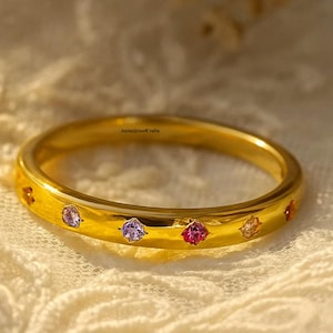 May include: A gold-colored ring with several small, colorful gemstones. The ring is set with purple, pink, and yellow stones. The ring is on a white lace surface.