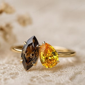 May include: A gold ring featuring two gemstones: a marquise-cut brown stone and a pear-shaped yellow stone. The ring is set against a soft, textured background, highlighting the sparkle of the gems.