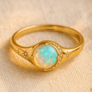 Op de afbeelding: Een gouden ring met een ovale opaal edelsteen. De ring heeft een halo van kleine diamanten rond de opaal en sterrenpatronen op de band. De opaal heeft een kleurenspel met tinten blauw, groen en geel.