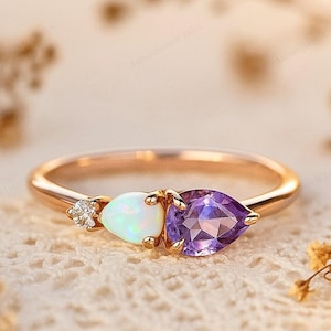 Könnte beinhalten: Ein goldener Ring mit einem kleinen Diamanten, einem Opal und einem birnenförmigen Amethysten. Der Ring liegt auf einer strukturierten, cremefarbenen Oberfläche, mit getrockneten Blumen im Hintergrund.
