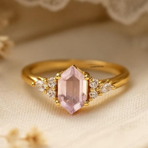 Op de afbeelding: Een gouden ring met een zeshoekige, lichtroze edelsteen in het midden. Kleine, heldere edelstenen zijn aan weerszijden van de hoofdsteen gezet. De ring wordt gepresenteerd tegen een zachte, neutrale achtergrond, die het ontwerp van de sieraden benadrukt.