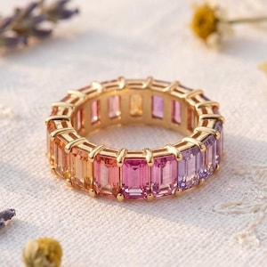 May include: A gold ring with a band of rectangular, multi-colored gemstones. The gemstones transition from yellow to pink to purple. The ring is set on a white lace surface, with dried flowers and petals scattered around it.