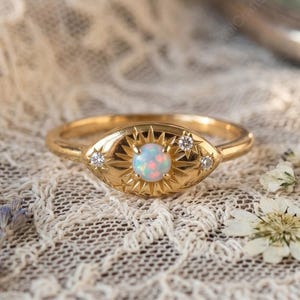 May include: A gold ring with an oval-shaped face featuring a central opal stone and starburst design. Two small clear stones flank the opal. The ring is set on a white lace surface.