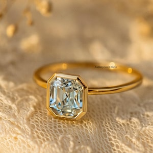 May include: A gold ring featuring a large, rectangular, pale blue gemstone. The gemstone is set in a gold bezel. The ring rests on a cream-coloured, lace-like fabric. The text "AuranjewelCrafts" is visible on the ring.