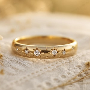 Puede incluir: Un anillo dorado con una serie de diseños en forma de estrella, cada uno con una pequeña gema transparente. El anillo se encuentra sobre un fondo suave y borroso, resaltando los intrincados detalles de la joya.