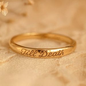 May include: A gold-colored ring with the inscription "Till Death" is displayed on a textured, cream-colored surface. The ring is the focal point, with a shallow depth of field. Delicate, dried flowers are in the background.
