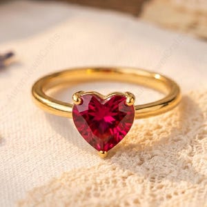 Puede incluir: Un anillo de oro con una piedra preciosa roja oscura en forma de corazón. La banda del anillo es de oro pulido, y la piedra preciosa está sujeta por cuatro garras. El anillo está sobre un fondo suave y texturizado.