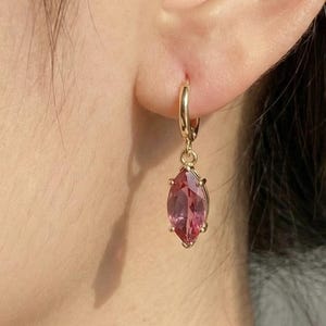 Puede incluir: Pendientes de aro dorados con un colgante de piedra preciosa rosa con forma de marquesa. Los pendientes se muestran en la oreja de una persona, destacando el diseño y el color de la joya. La piedra preciosa es de un tono rosa vibrante.