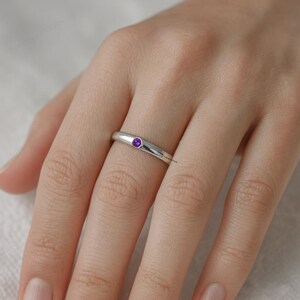 May include: A silver ring with a small, round, purple gemstone set in the center. The ring is smooth and polished, reflecting light. The gemstone is a vibrant purple color, adding a pop of color to the minimalist design. The ring is worn on a finger.
