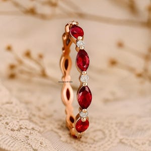 Könnte beinhalten: Roségoldener Ring mit abwechselnd oval geschliffenen roten Edelsteinen und kleinen, runden, klaren Edelsteinen. Der Ring ist vor einem weichen, neutralen Hintergrund platziert, der sein elegantes Design hervorhebt. Der Ring ist ein Schmuckstück.