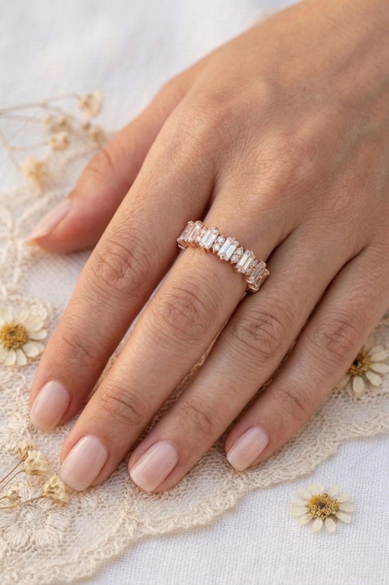 Op de afbeelding: Een ros&eacute;gouden ring met een reeks rechthoekige en rond geslepen heldere edelstenen. De ring zit aan een hand met neutrale nagellak. De achtergrond omvat kant en gedroogde bloemen.