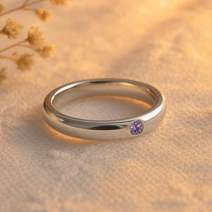 May include: A silver ring with a single, round, purple gemstone. The ring is set against a soft, textured background with a sprig of dried flowers in the upper left corner. The ring's band is smooth and polished.