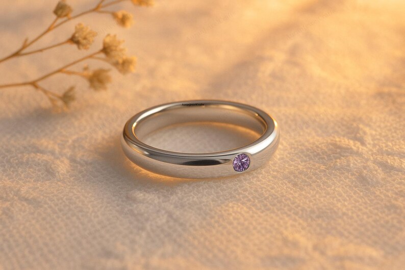 May include: A silver ring with a single, round, purple gemstone. The ring is set against a soft, textured background with a sprig of dried flowers in the upper left corner. The ring's band is smooth and polished.