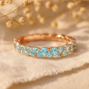 Könnte beinhalten: Ein roségoldener Ring mit einem Band aus hellblauen Edelsteinen. Die Edelsteine sind in verschiedenen Formen und Größen geschliffen, wodurch ein funkelndes Design entsteht. Der Ring liegt auf einem neutralfarbenen Stoff.