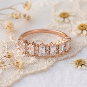 Op de afbeelding: Ros&eacute;gouden ring met een band van rechthoekige en rond geslepen heldere edelstenen. De ring ligt op een wit kanten oppervlak met gedroogde bloemen.