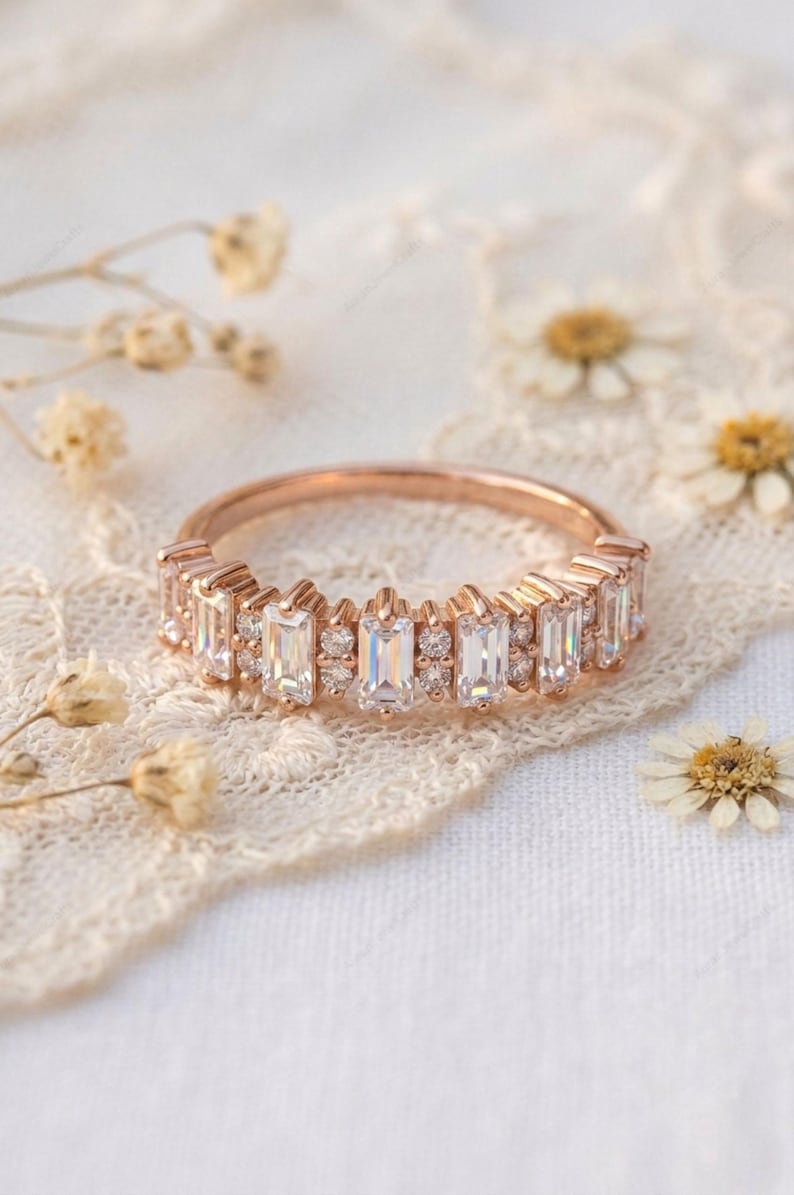 Op de afbeelding: Ros&eacute;gouden ring met een band van rechthoekige en rond geslepen heldere edelstenen. De ring ligt op een wit kanten oppervlak met gedroogde bloemen.