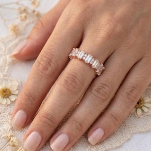 Op de afbeelding: Een ros&eacute;gouden ring met een reeks rechthoekige en rond geslepen heldere edelstenen. De ring zit aan een hand met neutrale nagellak. De achtergrond omvat kant en gedroogde bloemen.