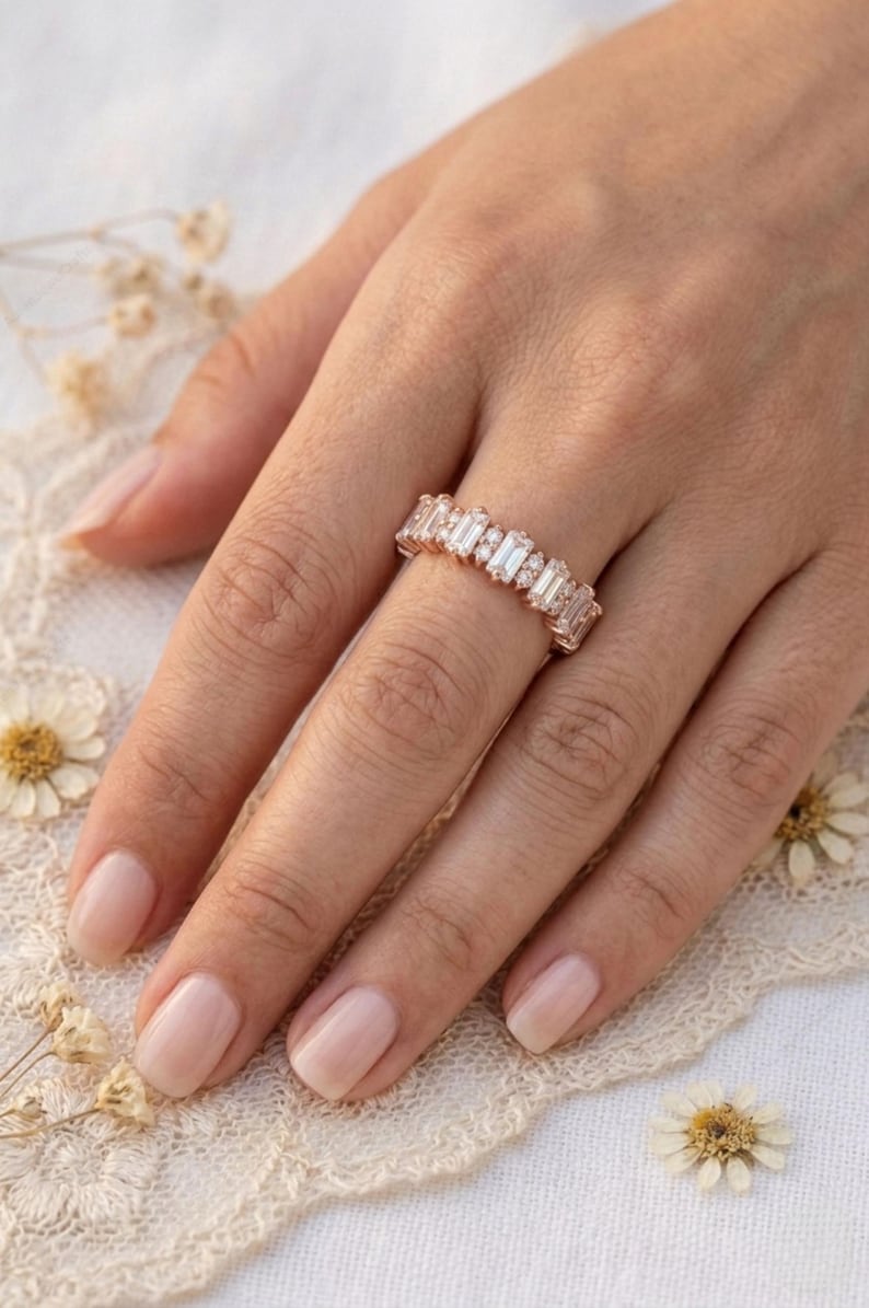 Op de afbeelding: Een ros&eacute;gouden ring met een reeks rechthoekige en rond geslepen heldere edelstenen. De ring zit aan een hand met neutrale nagellak. De achtergrond omvat kant en gedroogde bloemen.