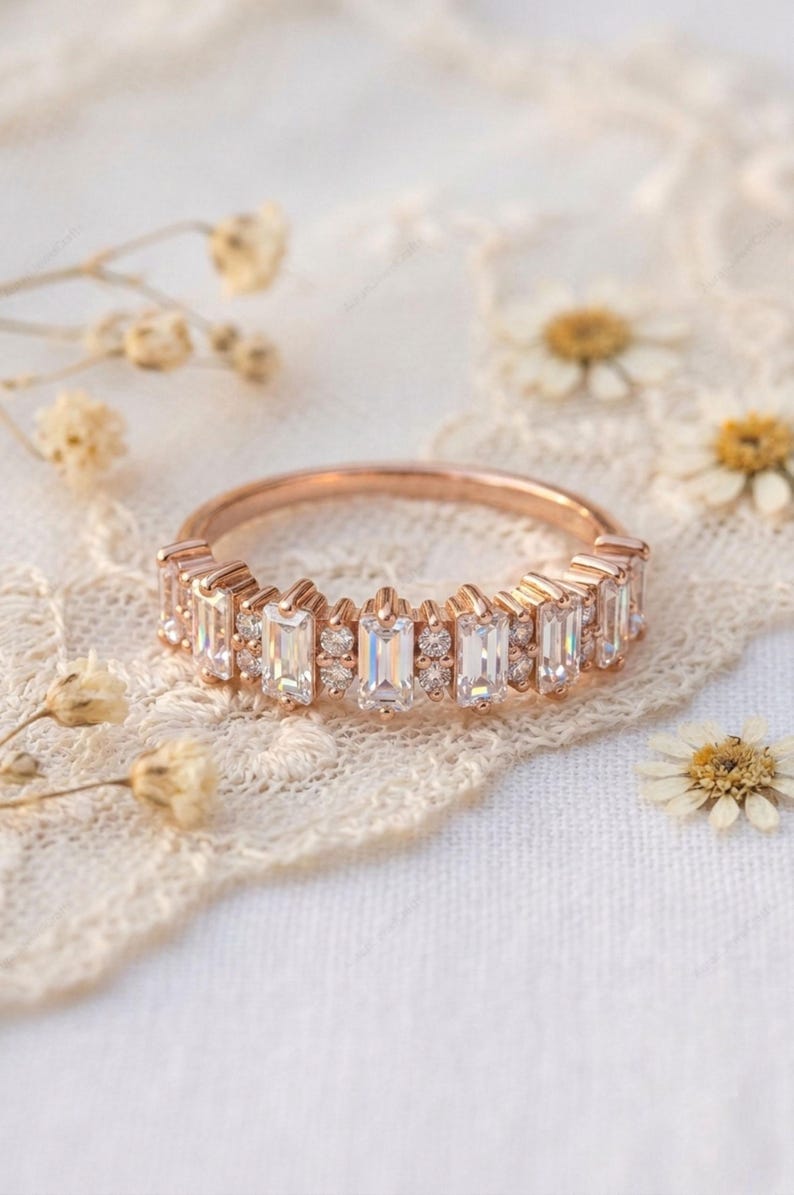 Op de afbeelding: Ros&eacute;gouden ring met een band van rechthoekige en rond geslepen heldere edelstenen. De ring ligt op een wit kanten oppervlak met kleine gedroogde bloemen.