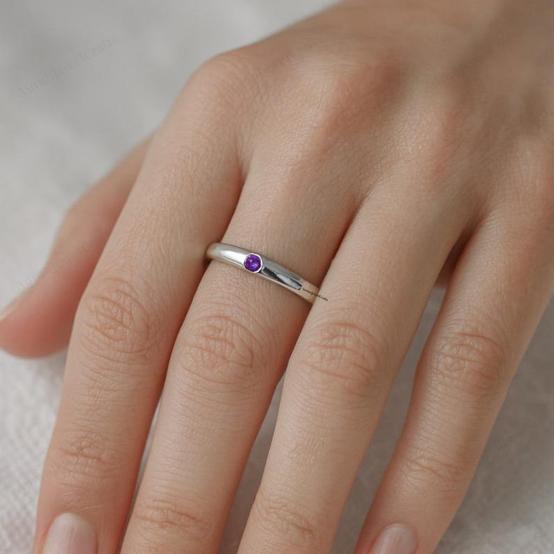 May include: A silver ring with a small, round, purple gemstone set in the center. The ring is smooth and polished, reflecting light. The gemstone is a vibrant purple color, adding a pop of color to the minimalist design. The ring is worn on a finger.