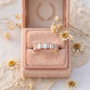 Op de afbeelding: Een gouden ring met rechthoekige en ronde edelstenen ligt in een roze fluwelen ringdoosje. Het doosje is open en onthult een cr&egrave;mekleurig interieur met een gouden lauwerkransontwerp. Gedroogde bloemen en kant omringen het doosje.