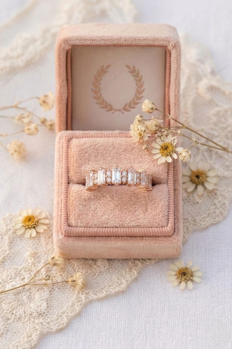 Op de afbeelding: Een gouden ring met rechthoekige en ronde edelstenen ligt in een roze fluwelen ringdoosje. Het doosje is open en onthult een cr&egrave;mekleurig interieur met een gouden lauwerkransontwerp. Gedroogde bloemen en kant omringen het doosje.