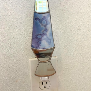 May include: A stained glass night light with a blue and purple cloud-like design. The lamp has a mirrored base and is plugged into a white wall outlet. The top of the lamp has a window-like design.