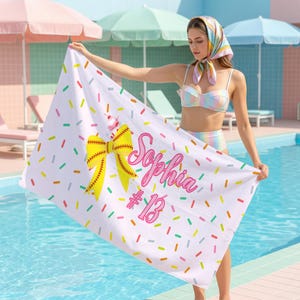 May include: White rectangular beach towel with a colourful sprinkle pattern and a softball theme. It features a yellow bow, a softball, and the name "Sophia" in pink with the number "13". The towel appears to be made of soft material.