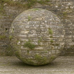 May include: A large, spherical object made of weathered, grey stone bricks. Green moss and grass grow between the bricks, adding texture and colour. The sphere rests on a weathered stone surface, with a brick wall in the background.