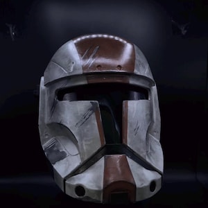 May include: A full-face helmet featuring a weathered white and brown color scheme. The design includes a horizontal visor opening, a central vertical ridge, and various simulated battle-worn scratches and scuffs across the surface.