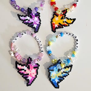 Puede incluir: Cuatro pulseras de cuentas con dijes pixelados en forma de corazón. Las pulseras presentan varios colores, incluyendo morado, rosa, amarillo y azul, con cuentas de letras blancas que deletrean frases como "WITH ALL MY" y "HOLD ON TO YOU".